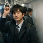 満員電車の車内でつり革を握る会社員。冬の朝、休み明けの「仕事始め」の疲れと再起動の表情。An office worker holding a train strap in a crowded commuter train on a winter morning, capturing the tired-but-determined “back to work” mood.