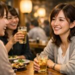 居酒屋のテーブルで、仕事終わりの女性たちがほろ酔いで笑い合いながら話を聞き合う瞬間。部署の人間関係が少しやさしく見えてくる空気感。 Three Japanese office women share drinks at an izakaya, leaning in with relaxed smiles, listening to each other in warm evening light that hints at gentle workplace camaraderie.